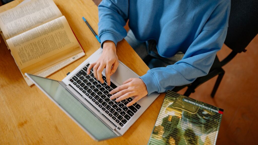 Student typing on a budget laptop with an open textbook and notebook