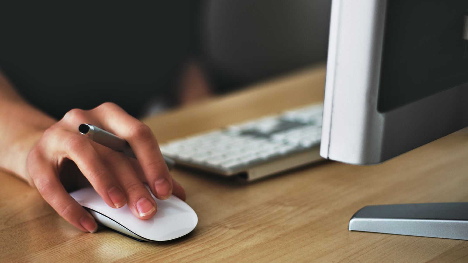 Hand resting on a wireless mouse at an office desk with keyboard and monitor