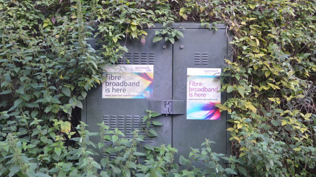 An Openreach fibre broadband street cabinet, half hidden by foliage, carrying "Fibre broadband is here" stickers.