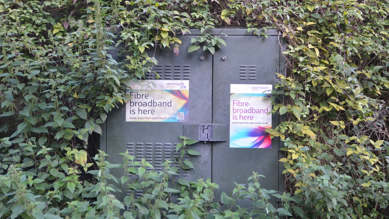 An Openreach fibre broadband street cabinet, half hidden by foliage, carrying "Fibre broadband is here" stickers.