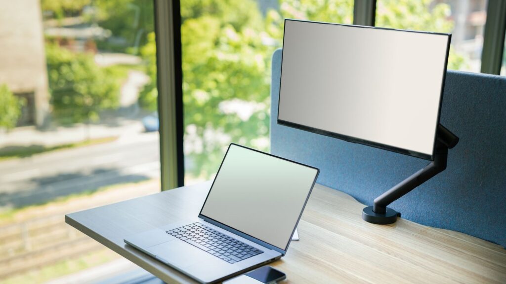 A computer monitor held on a black gas spring arm clamped to a desk, beside a laptop
