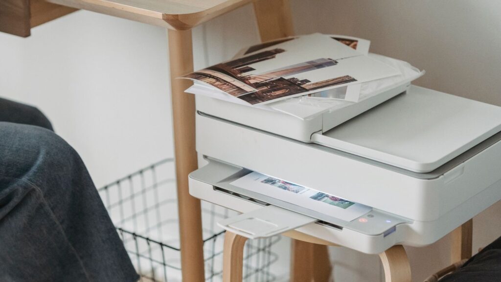 A white all-in-one inkjet printer on a stool beside a home desk, printing a page with photos stacked on its lid