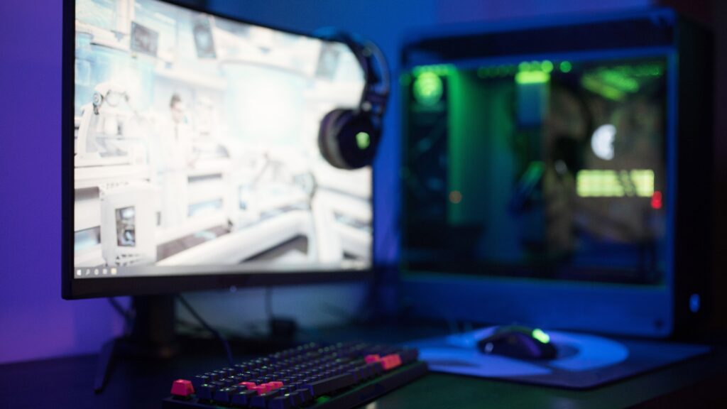 Curved gaming monitor next to a gaming PC with green lighting on a desk in a blue-lit room