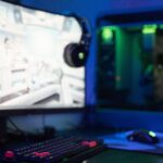 Curved gaming monitor next to a gaming PC with green lighting on a desk in a blue-lit room