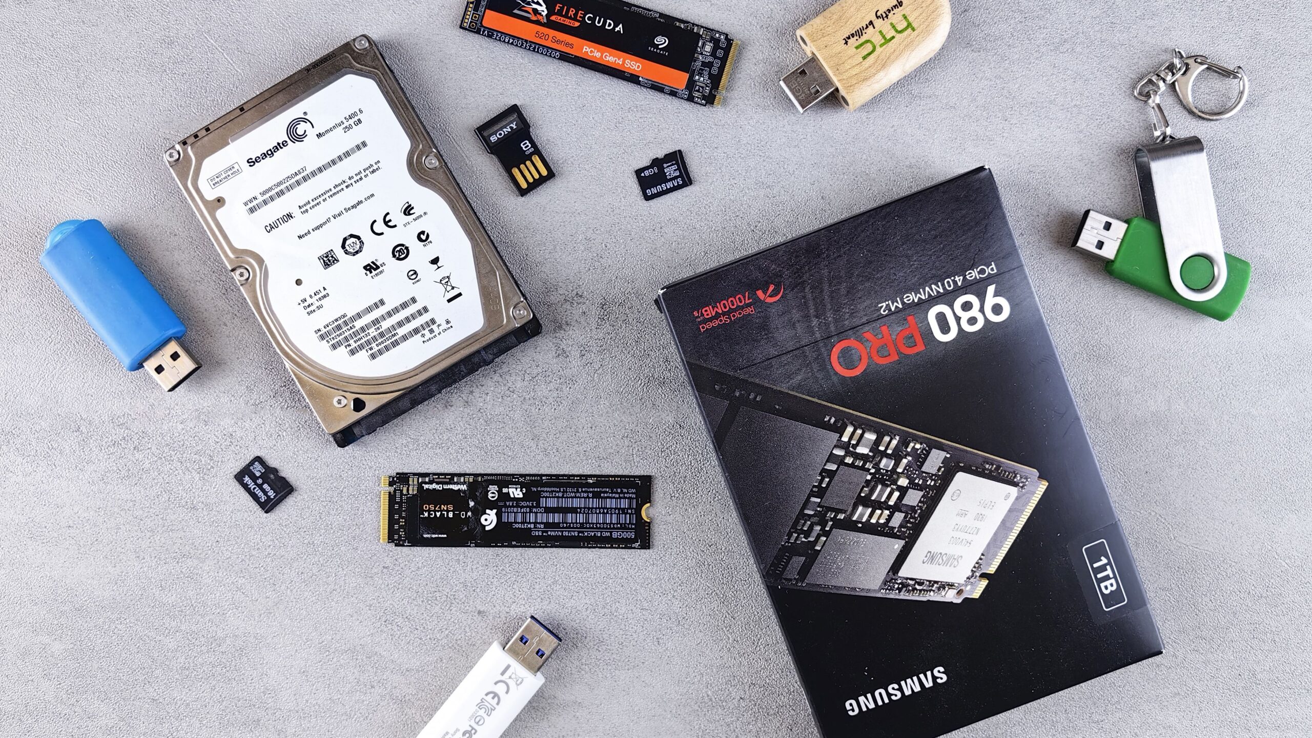M.2 NVMe SSDs, a laptop hard drive, USB sticks and memory cards laid out on a grey desk